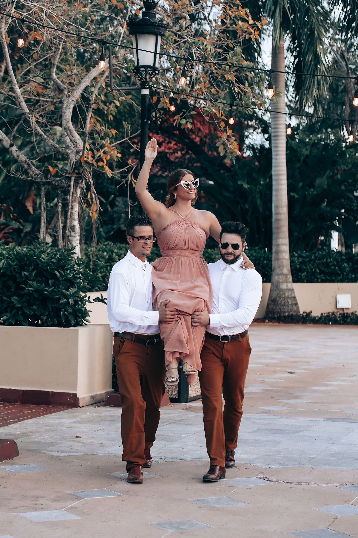 Three people in coordinated brown and white outfits pose playfully on a paved walkway near decorative street lamps and trees.