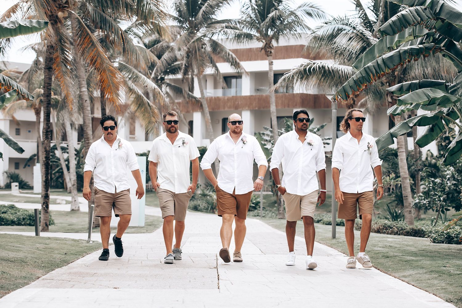 Wedding party walking along a palm-lined tropical beach path in coordinated white and beige outfits.