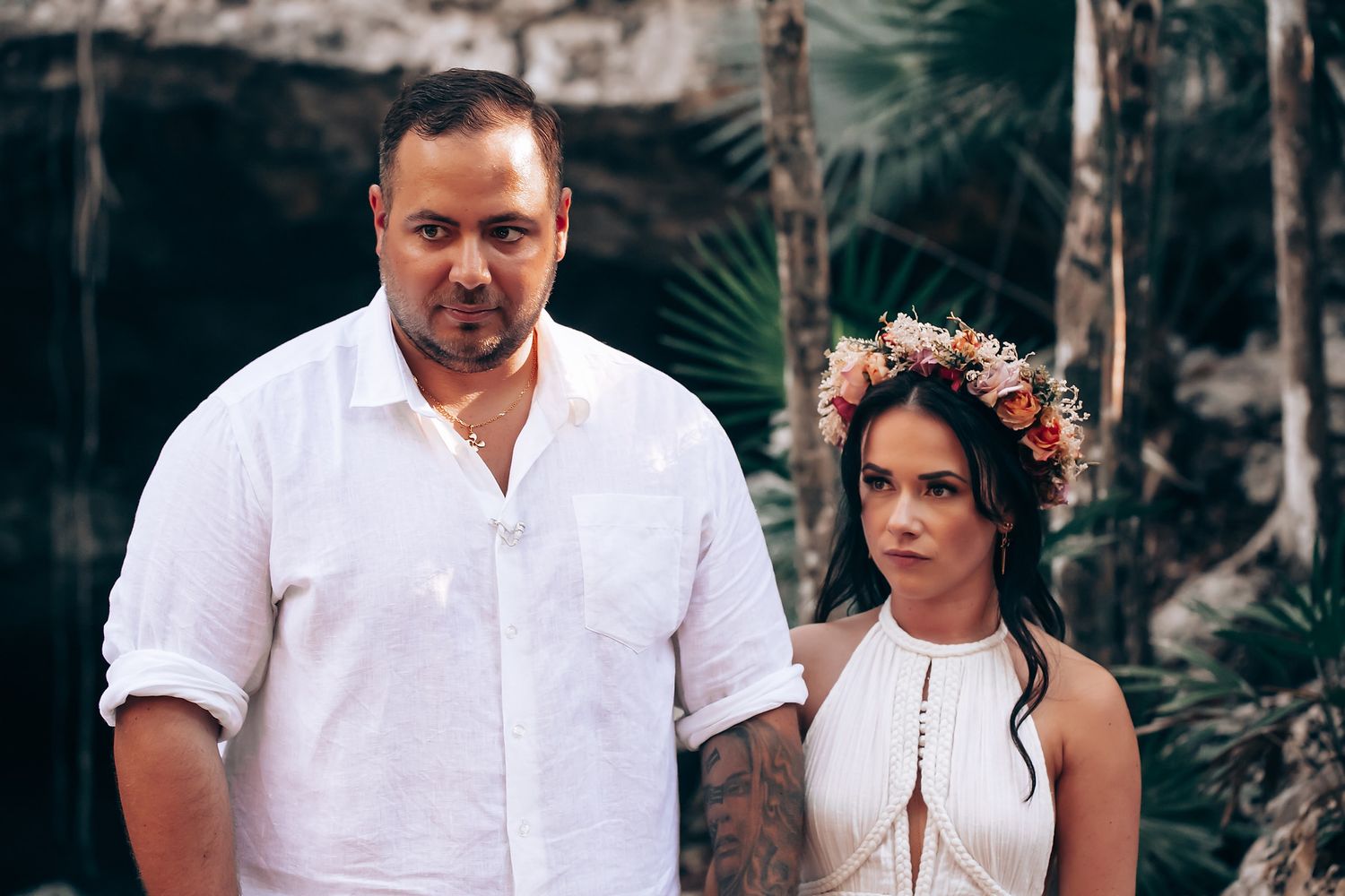 A couple in white attire poses together in a tropical outdoor setting surrounded by lush greenery.