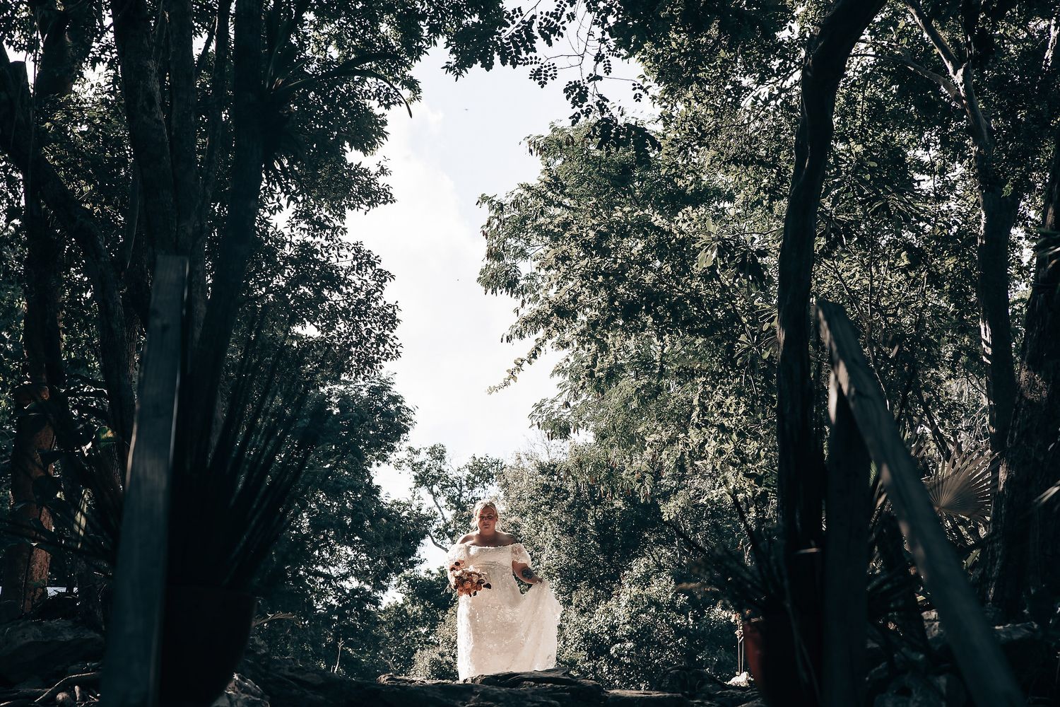 A woman in a white wedding dress standing on a wooden platform in a forest with trees and foliage in the background.