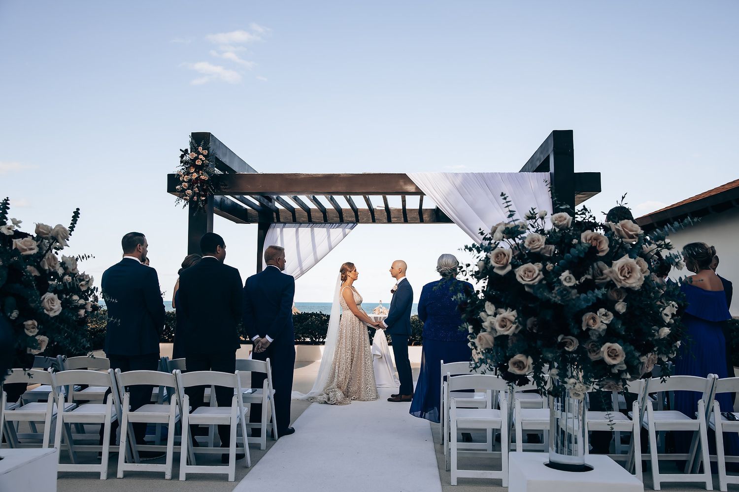A bride walks down an aisle lined with white chairs and floral arrangements at an outdoor wedding ceremony.