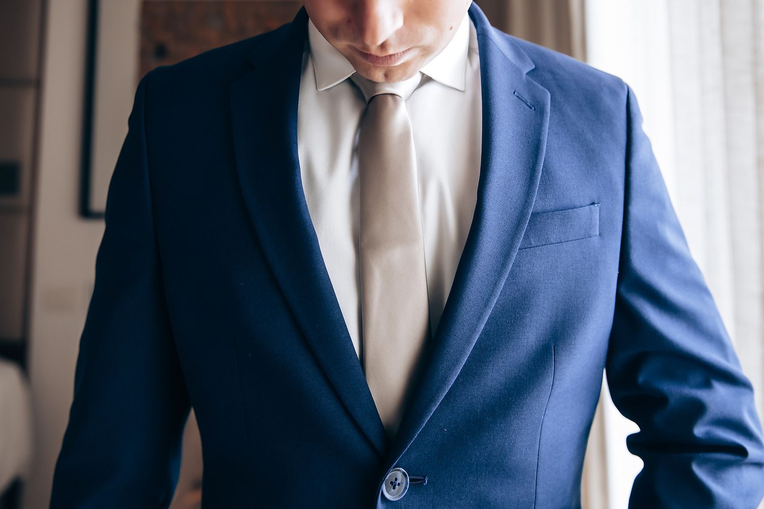 A man wearing a navy blue suit and tie stands indoors.
