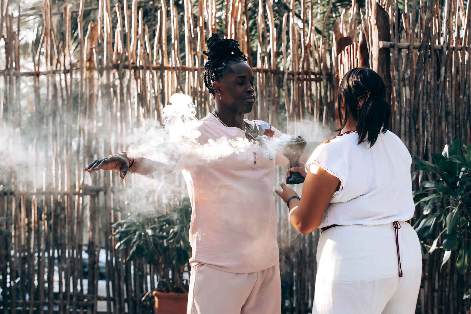 Two people, a man and a woman, stand in a tropical setting with palm trees and a thatched roof structure.
