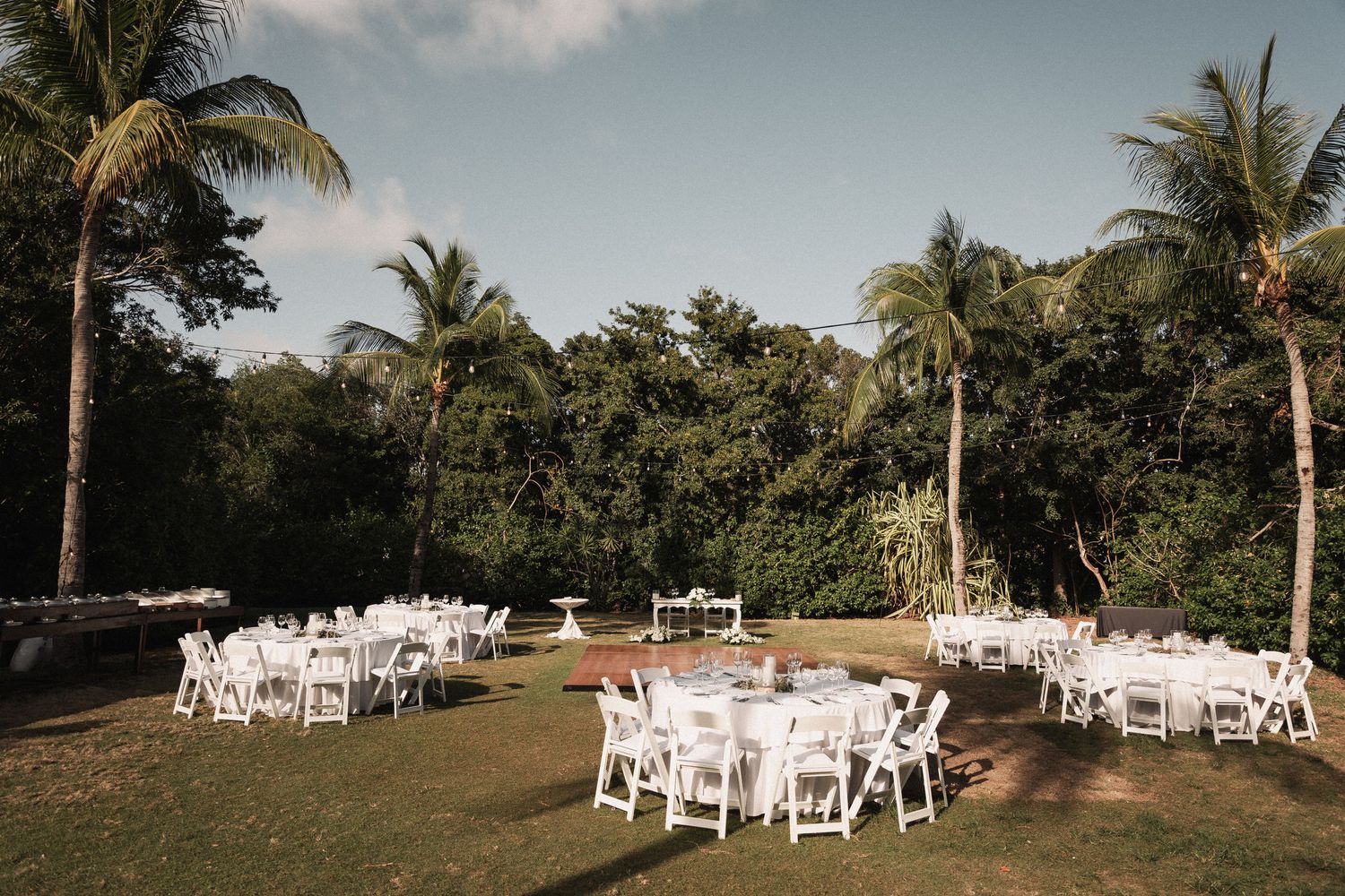 Outdoor tropical wedding reception setup with white round tables and chairs surrounded by palm trees.