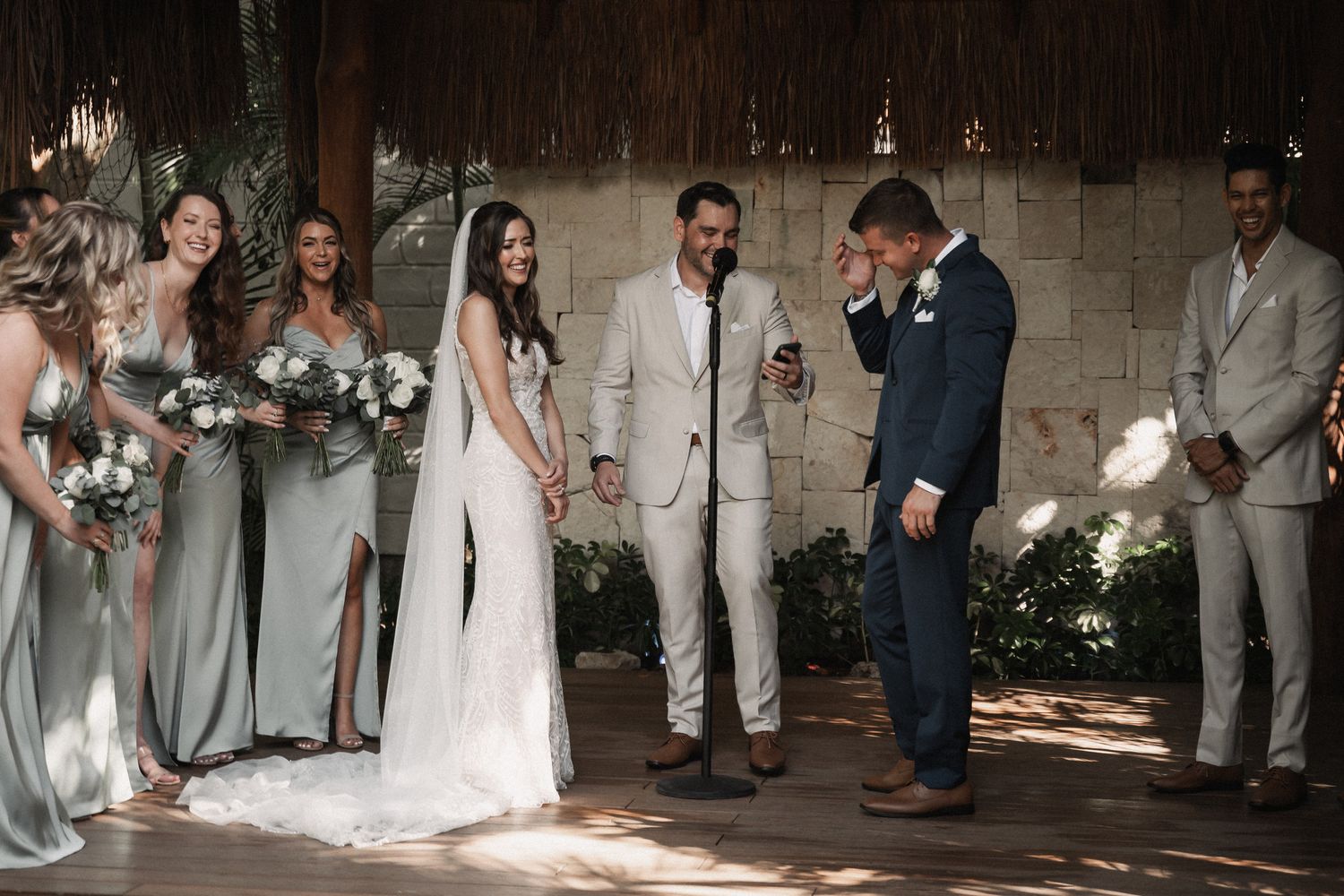 Bride and groom laugh together during outdoor wedding ceremony with bridesmaids in sage green dresses.