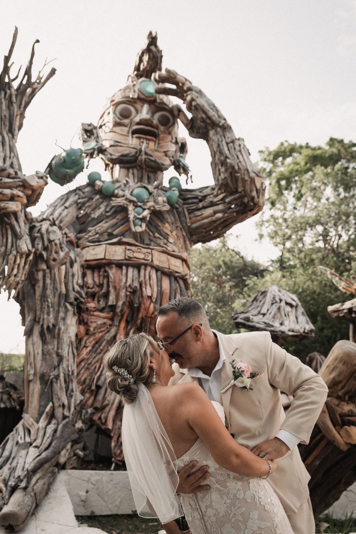 Bride and groom sharing a kiss in front of a large rustic driftwood sculpture at their wedding.