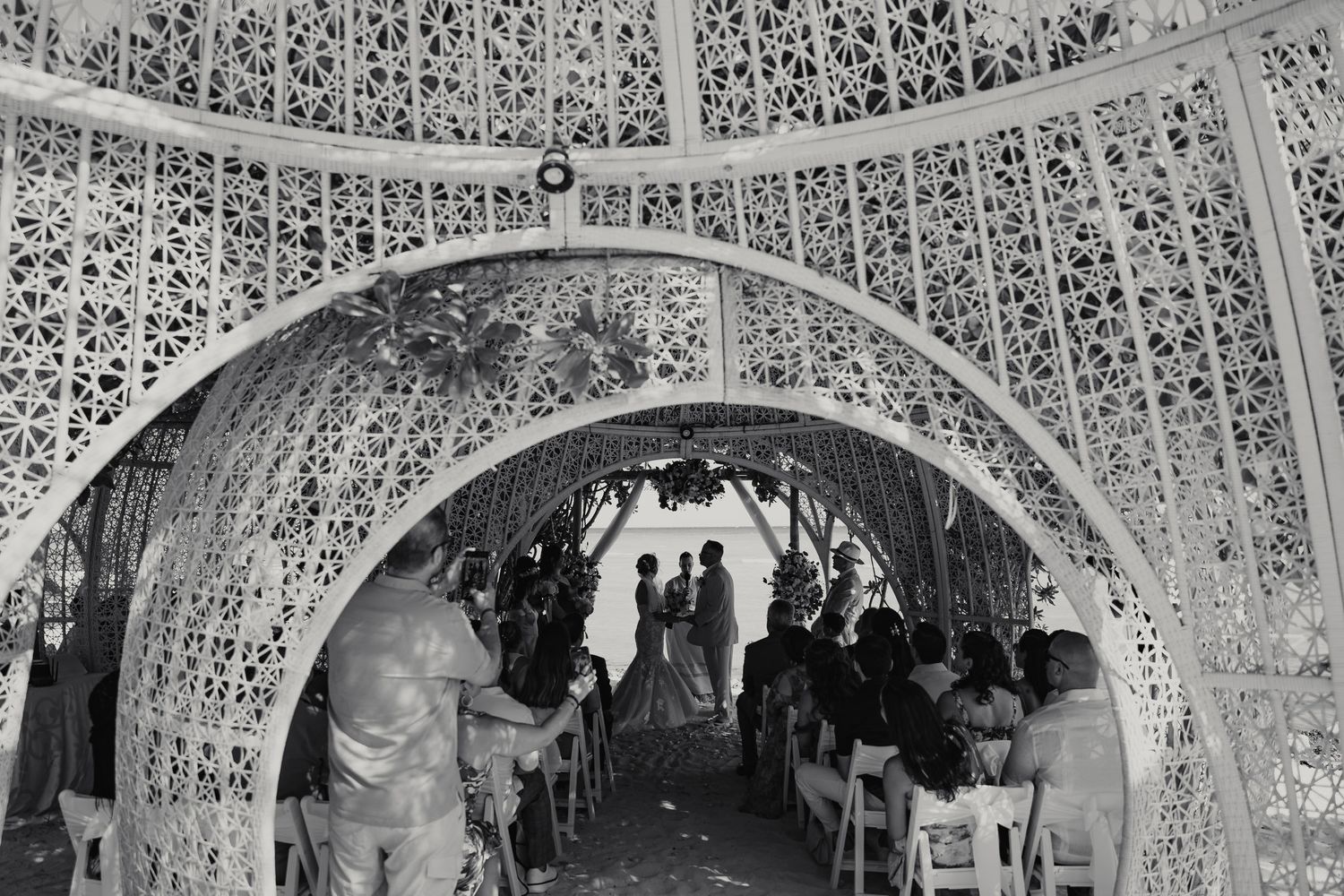Black and white photo of wedding ceremony under ornate white lattice arch with seated guests.