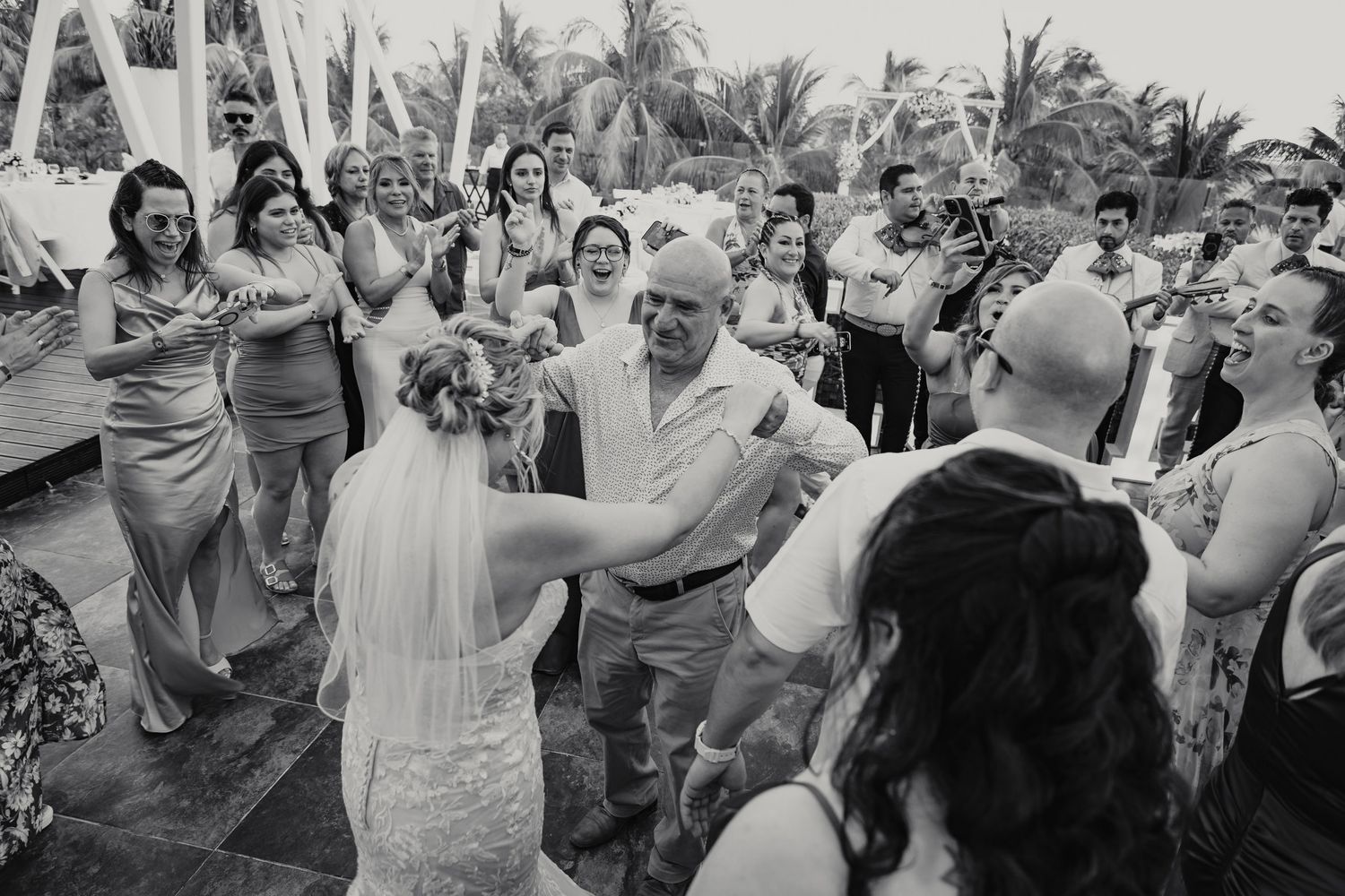 Black and white photo of lively outdoor wedding reception dance with guests and bride in veil.