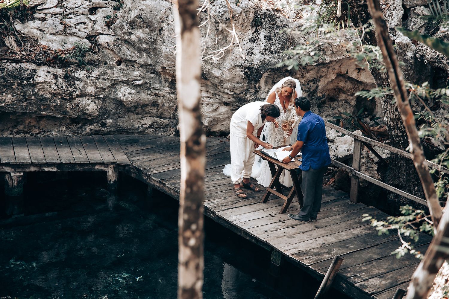 A couple sits together on a wooden dock over dark water surrounded by rocky cliffs.