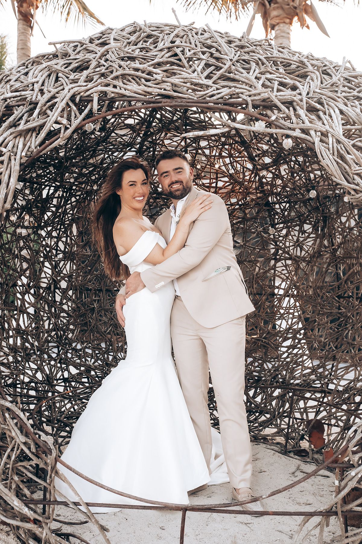 A couple in wedding attire poses together under a rustic wooden archway in a romantic outdoor setting.