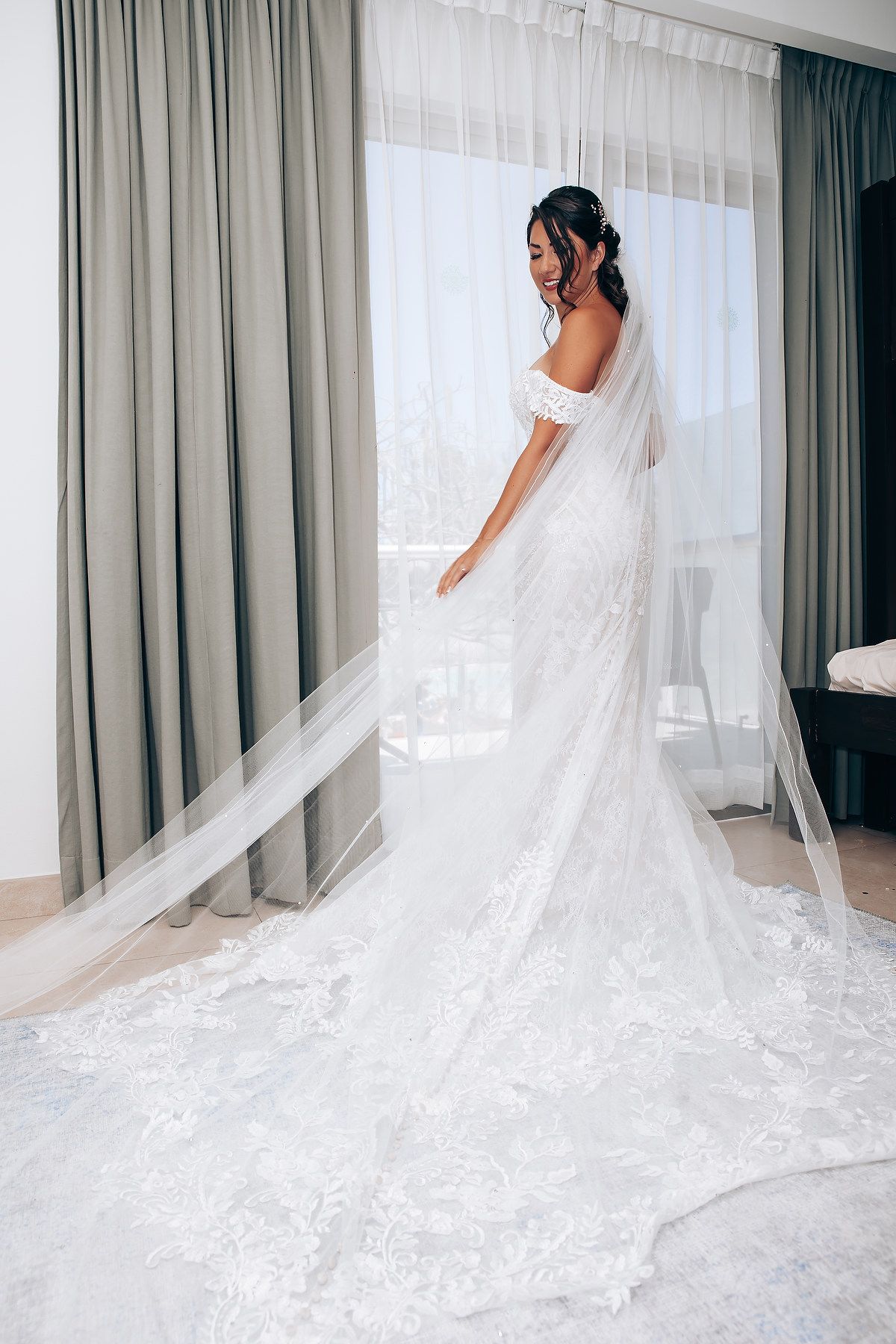 Elegant white wedding dress with flowing lace train displayed against window light in bridal suite.
