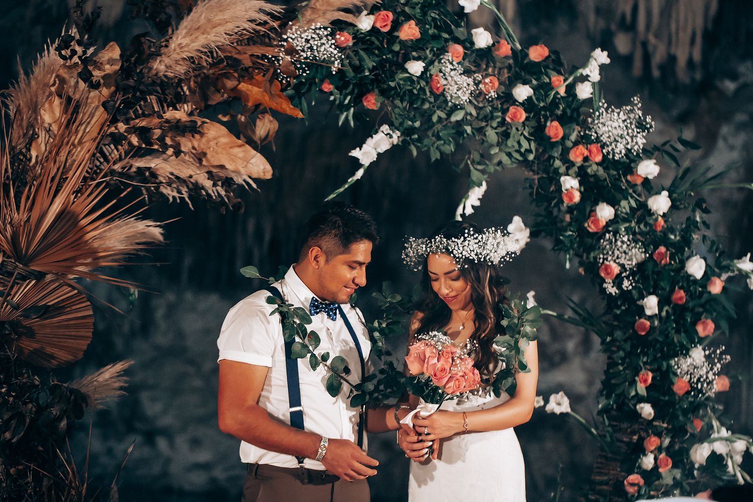 A romantic wedding couple shares an intimate moment surrounded by tropical flowers and palm fronds in a garden setting.