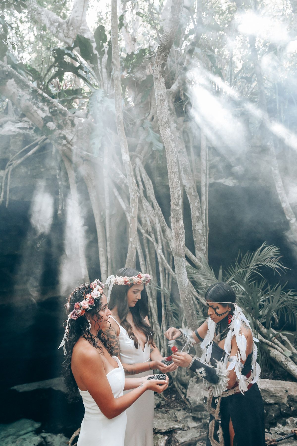 A couple in white dresses share an intimate moment in a tropical forest setting with dramatic rays of sunlight.