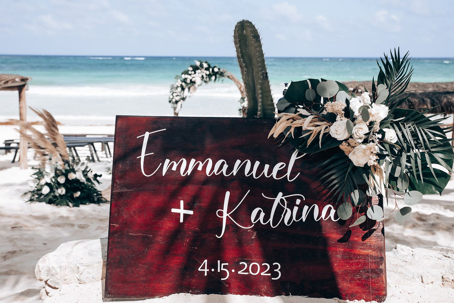 Wooden wedding welcome sign with names and date displayed on rustic red backdrop at beach venue.