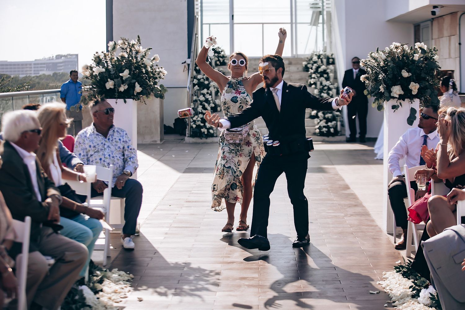 Wedding guests seated outdoors at a daytime ceremony with floral arrangements and white decor.