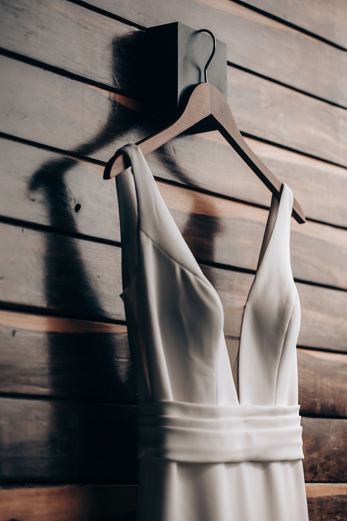 Elegant white wedding dress hanging against wooden slat wall in dramatic lighting.