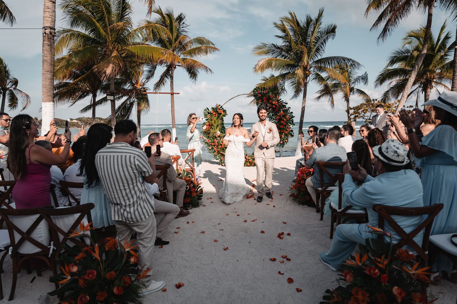 Newlyweds walk down sandy aisle as guests cheer at a tropical beach wedding ceremony at sunset.