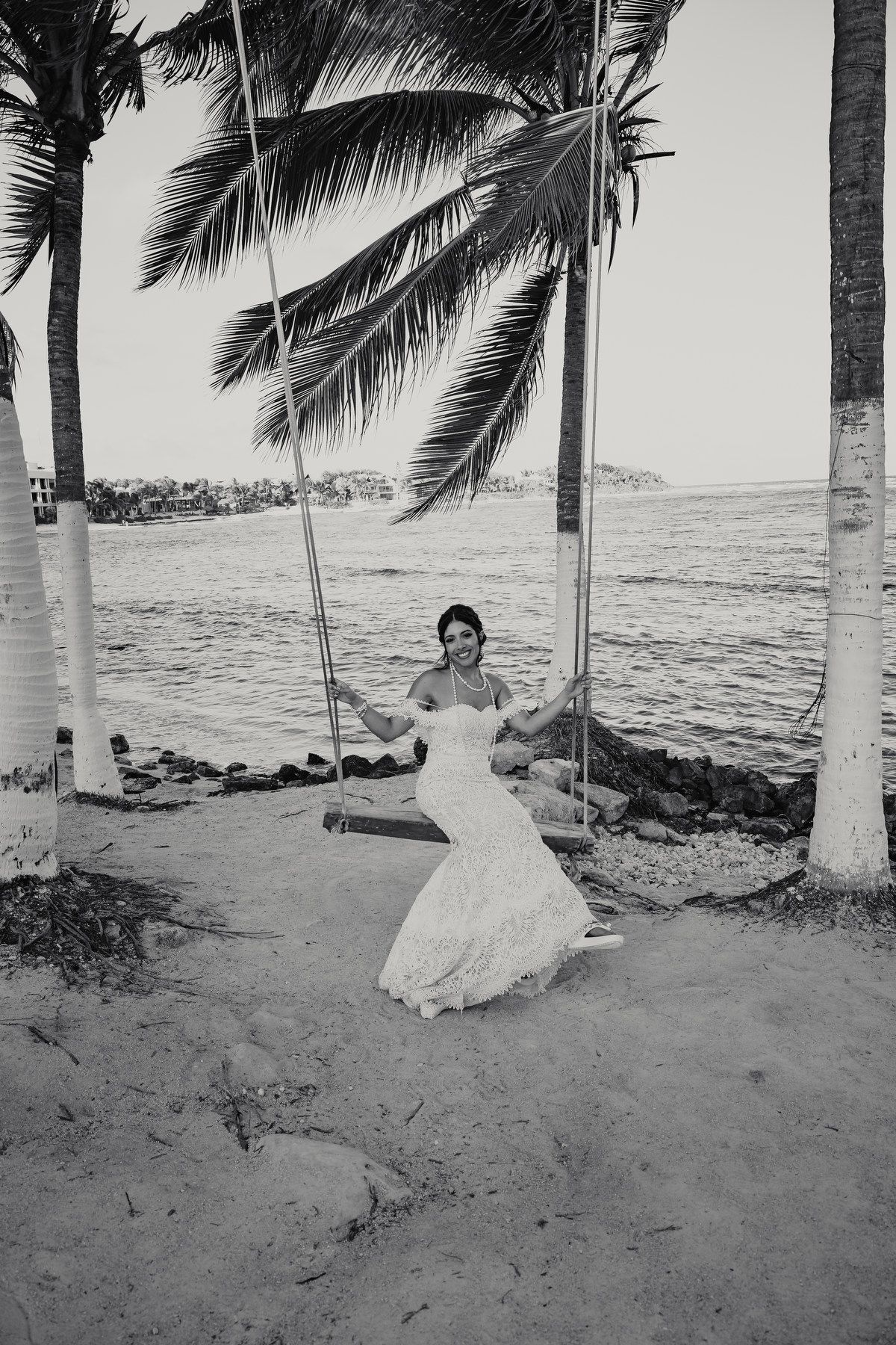 Black and white photo of bride in lace gown sitting on rope swing by palm trees.