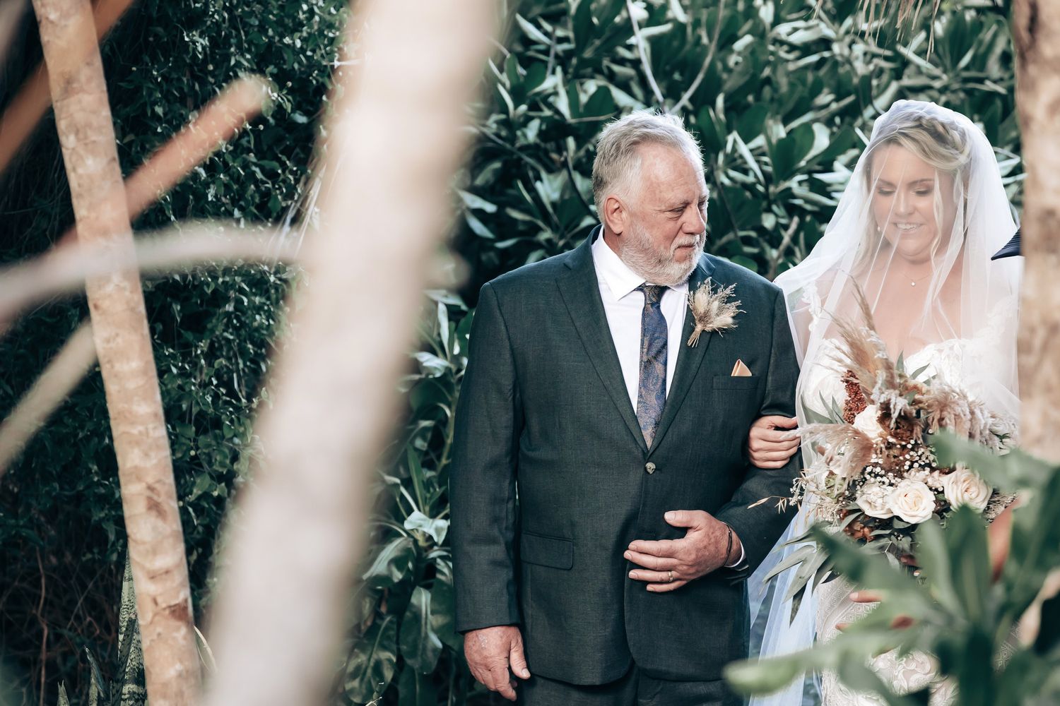 A bride in a white gown and veil walks with an older man in a suit through a lush garden ceremony.
