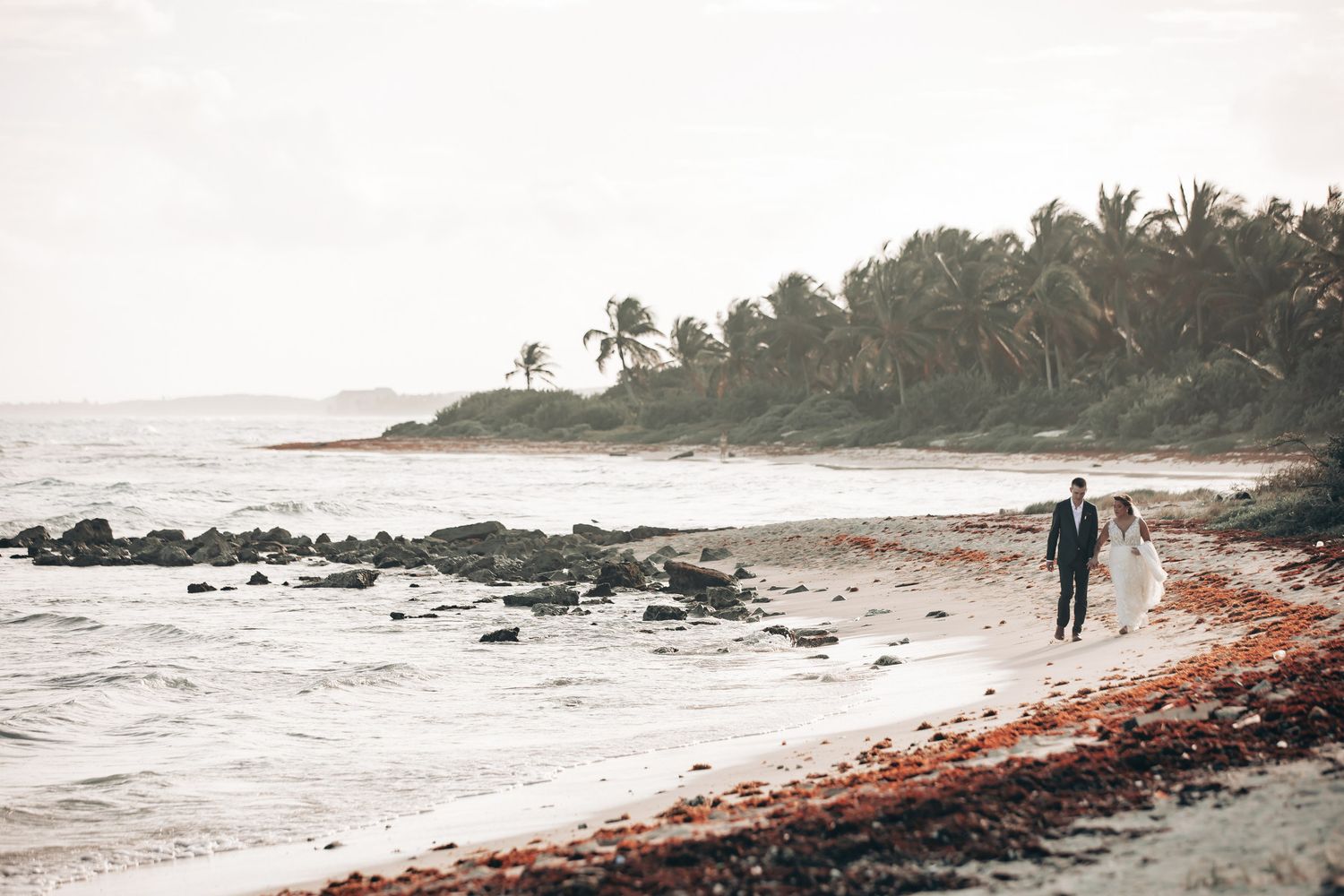 Bride and groom walking along a scenic tropical beach shoreline with palm trees in the background.