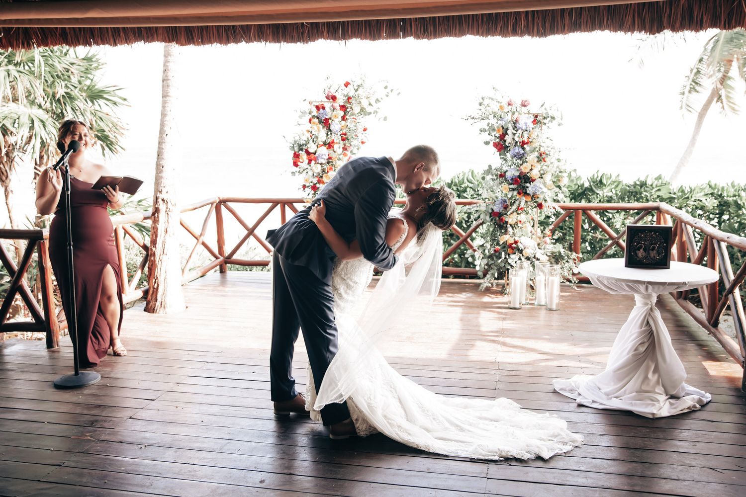 Groom dips bride in a romantic kiss at outdoor wedding ceremony with vibrant floral backdrop.