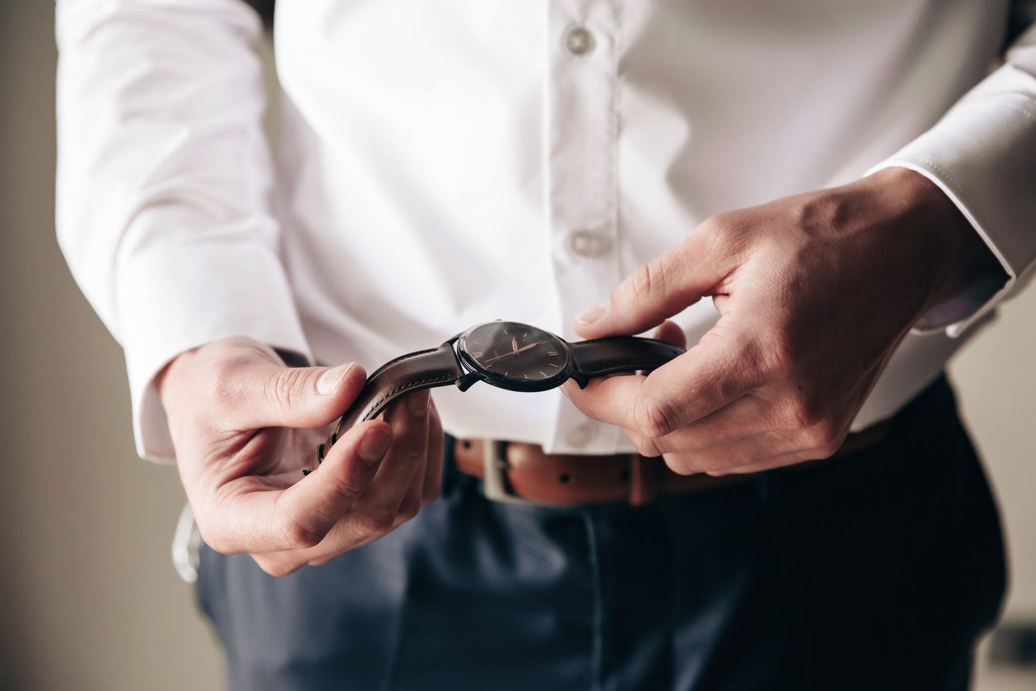 Groom in white dress shirt and navy pants putting on a black wristwatch with brown leather belt detail.