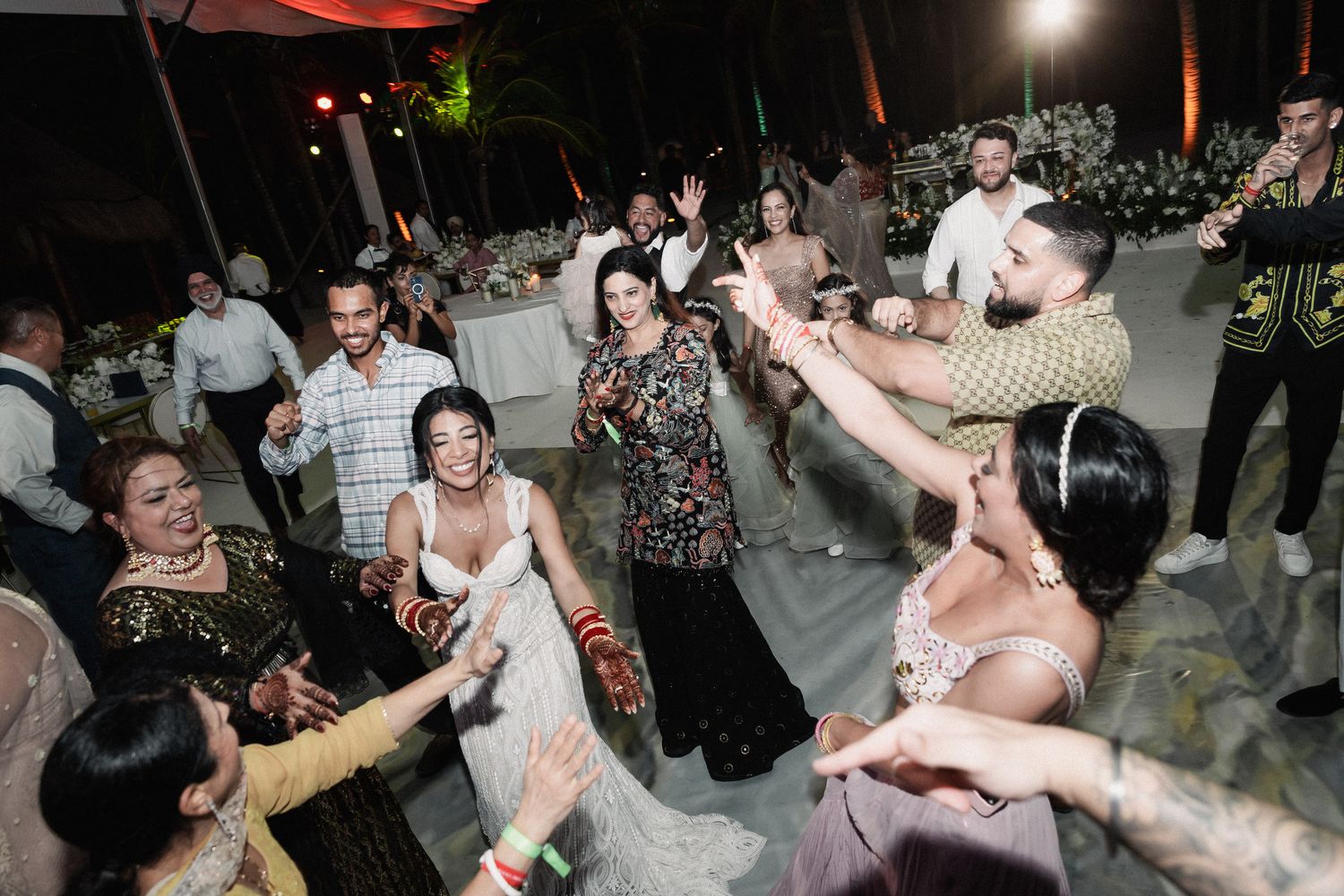 Lively wedding reception dance floor with guests celebrating around bride in white gown outdoors at night.