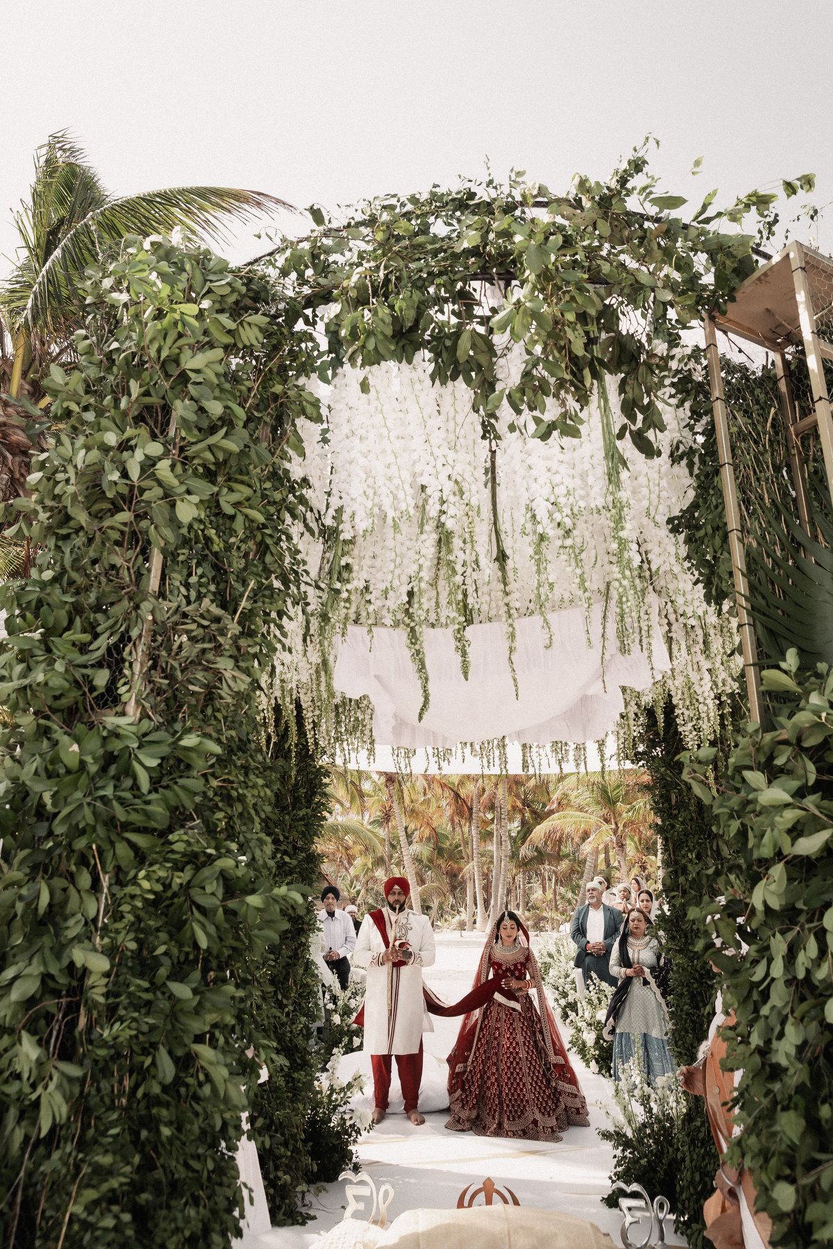 Indian bride and groom standing under lush greenery wedding arch adorned with hanging white flowers during beach ceremony.