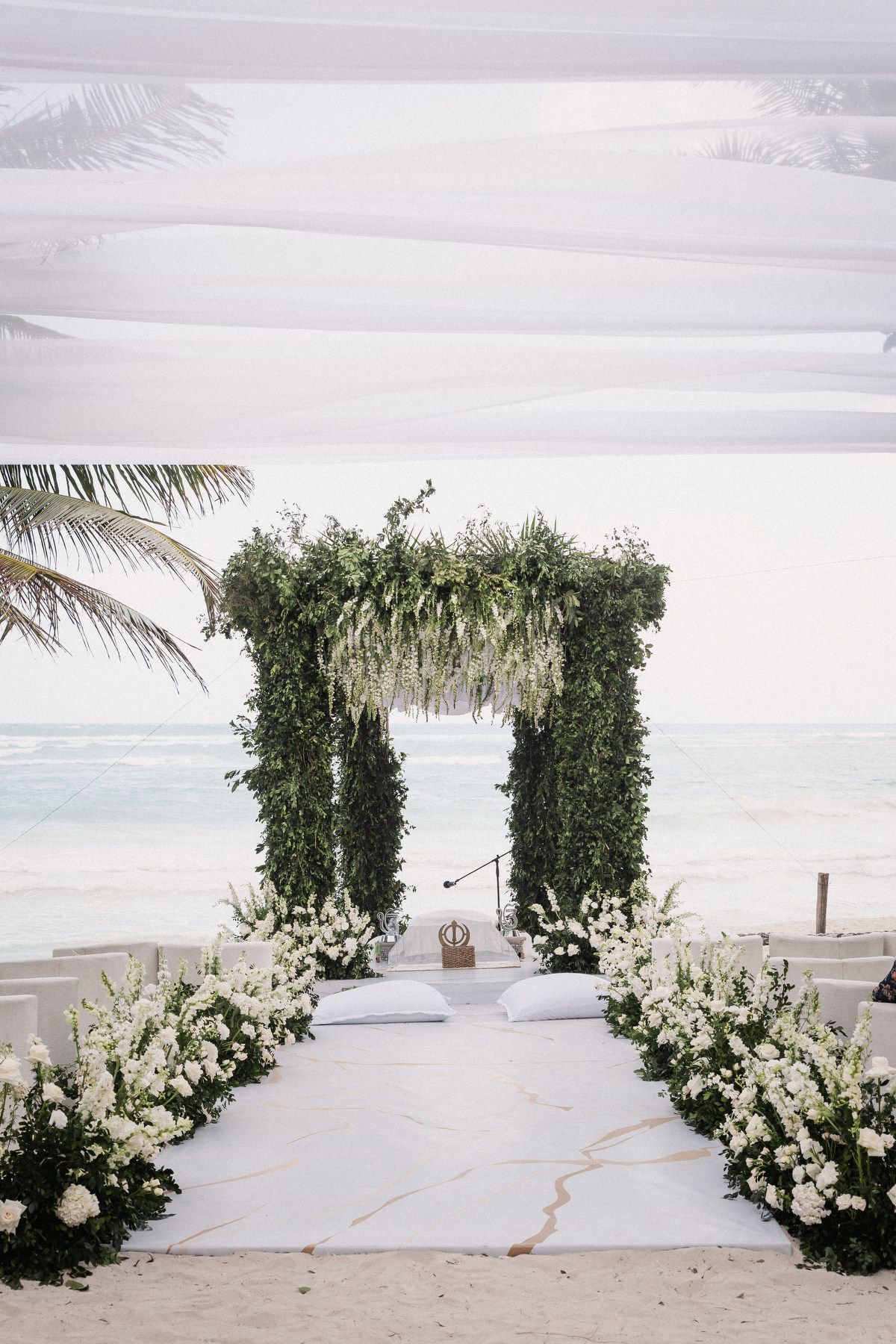 Lush green floral arch wedding ceremony setup on a beach with white flowers lining the aisle and ocean backdrop.