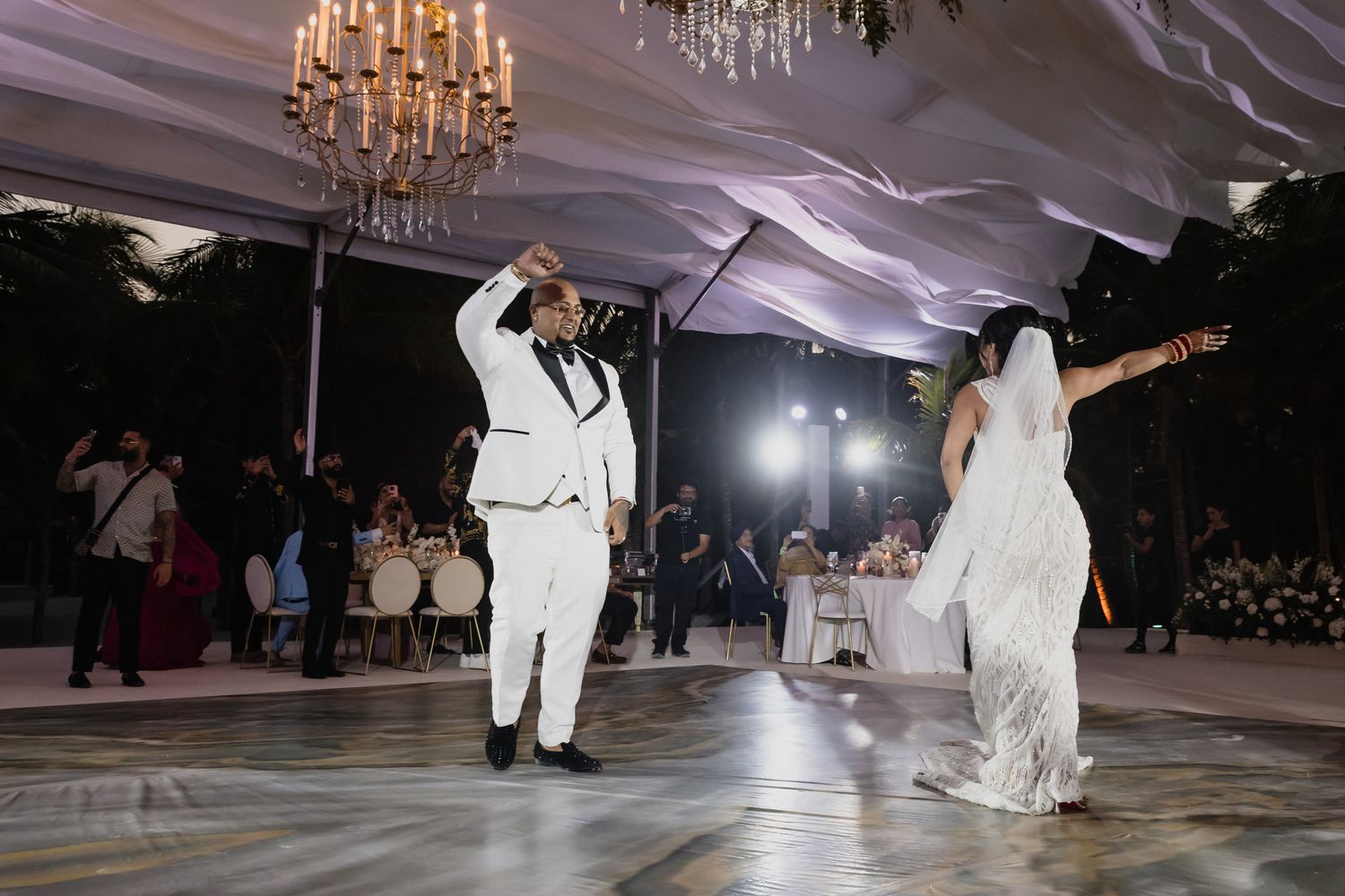 Bride and groom share an energetic first dance under a chandelier in an elegant outdoor wedding reception tent.