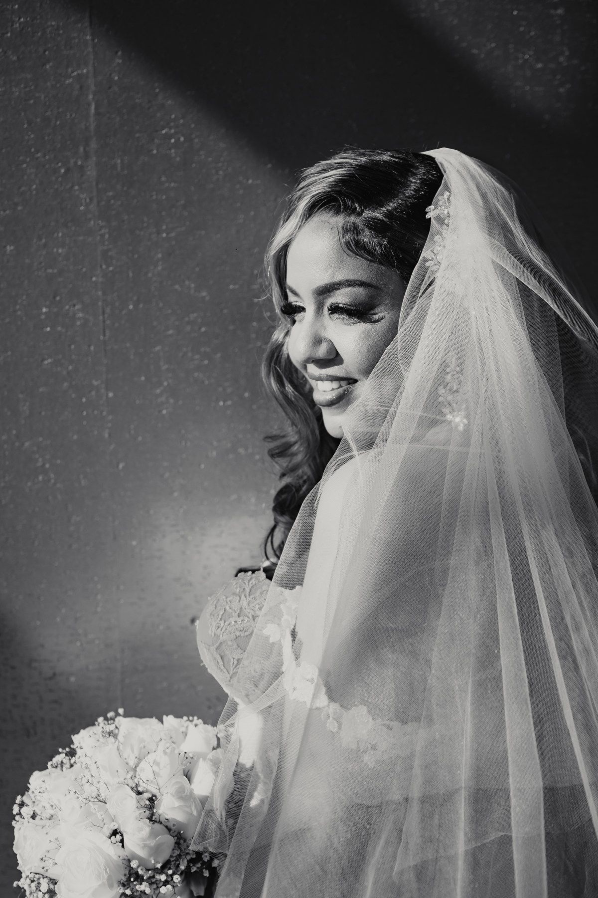 Black and white portrait of a smiling bride in veil holding a bouquet in soft dramatic lighting.
