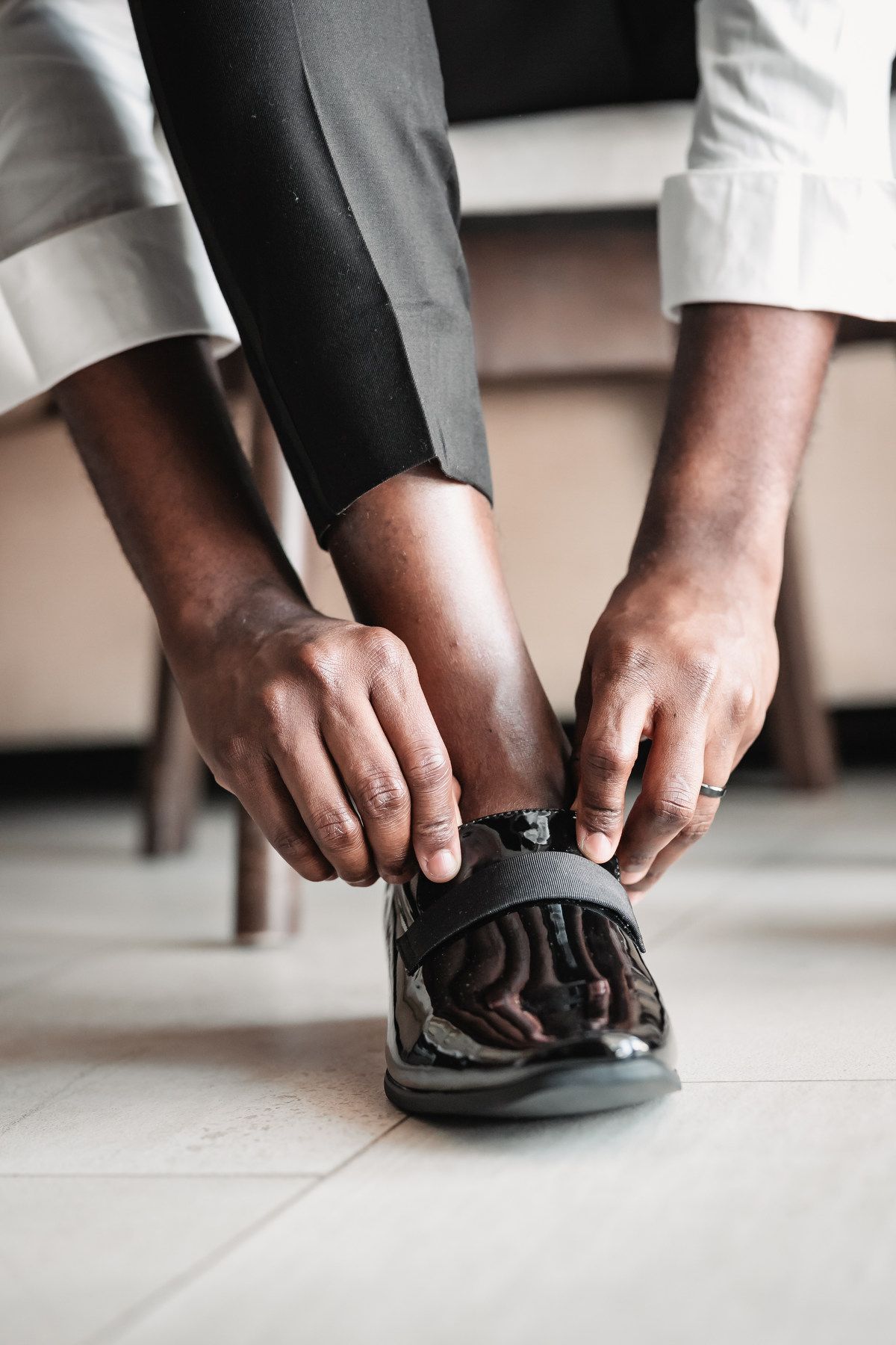 Groom fastening black patent leather dress shoes while wearing gray trousers on wedding day.