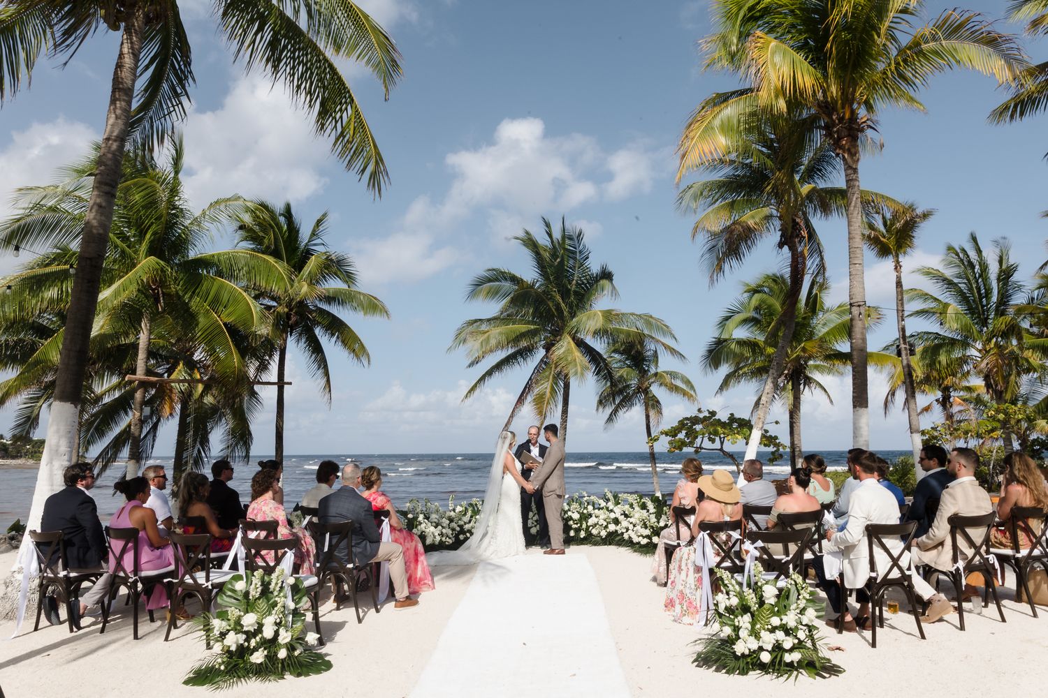 Outdoor beach wedding ceremony with guests seated among palm trees, white floral arrangements, and ocean views under a blue sky.