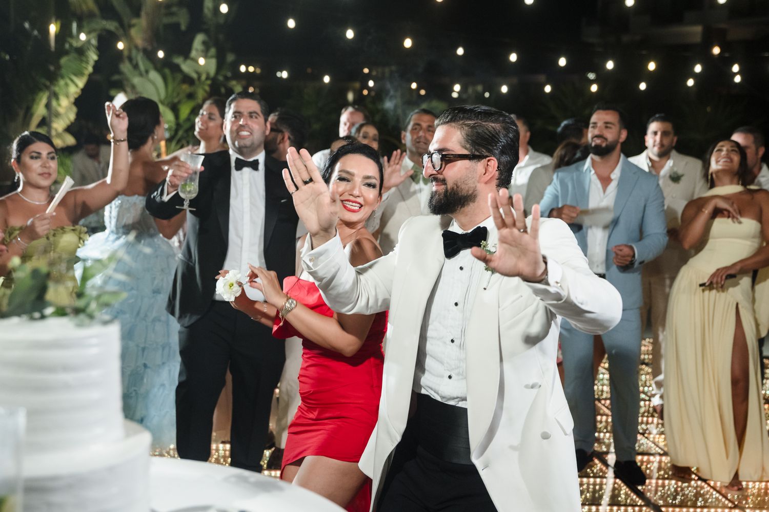 Bride in red dress and groom in white tuxedo dancing at outdoor wedding reception with guests applauding.