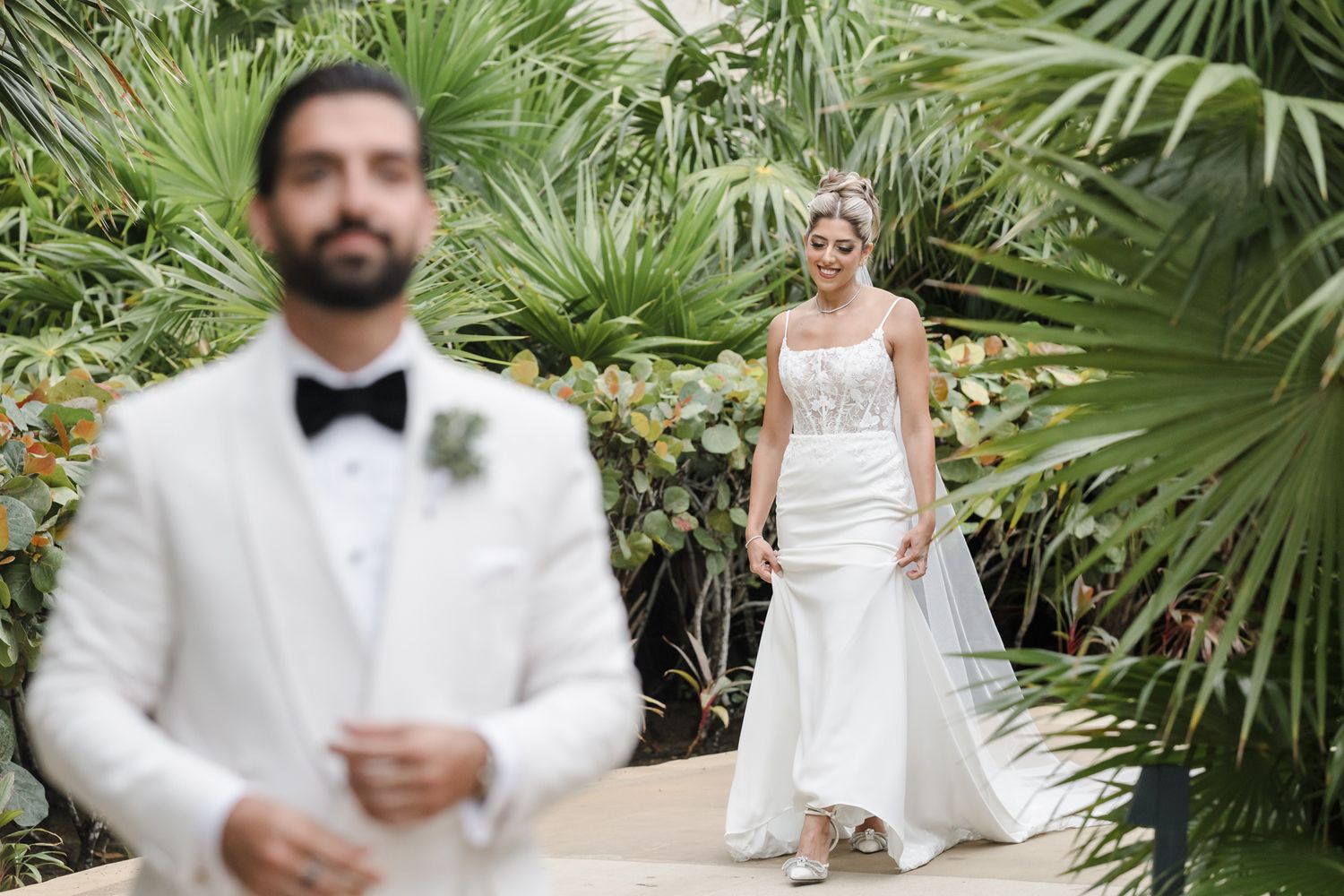 Bride in white gown approaches groom in white tuxedo during first look among lush tropical greenery.