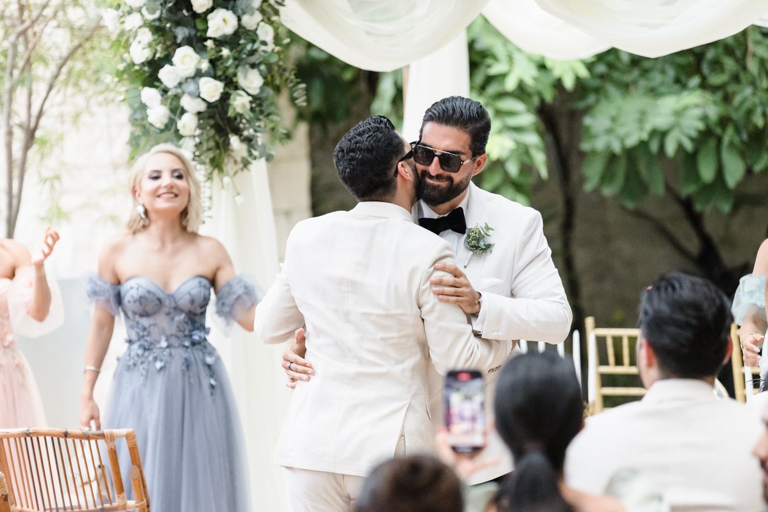 Two grooms embrace at outdoor wedding ceremony with floral arch, guests celebrating in elegant attire.
