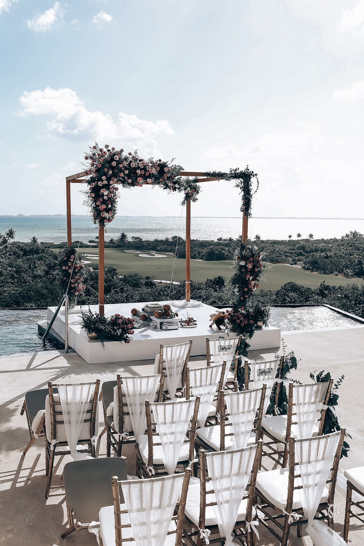 Elegant outdoor wedding ceremony setup with white chairs and floral arch overlooking the ocean.