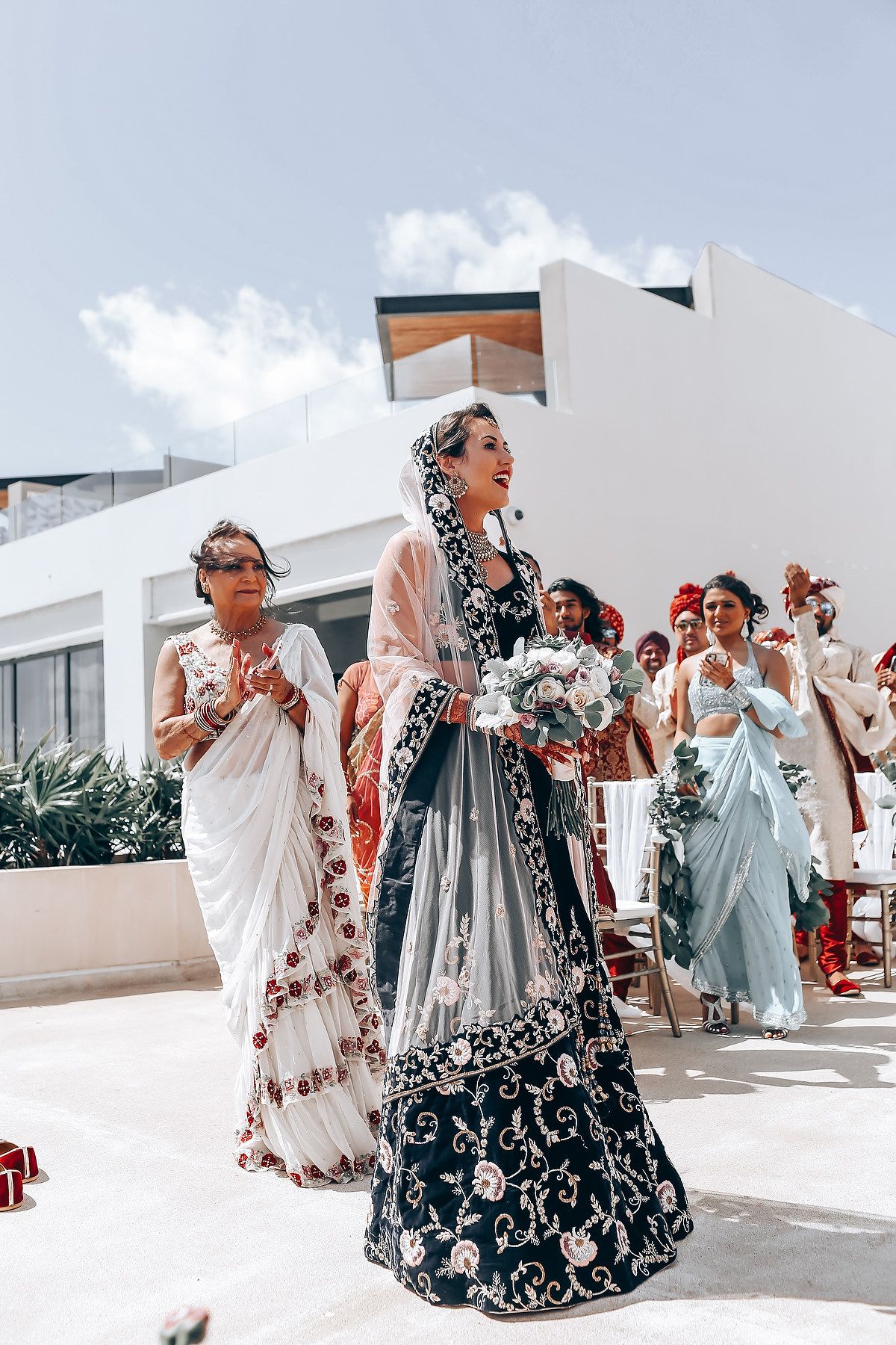 Elegant wedding guests in traditional Indian attire walk during an outdoor celebration with white buildings in background.
