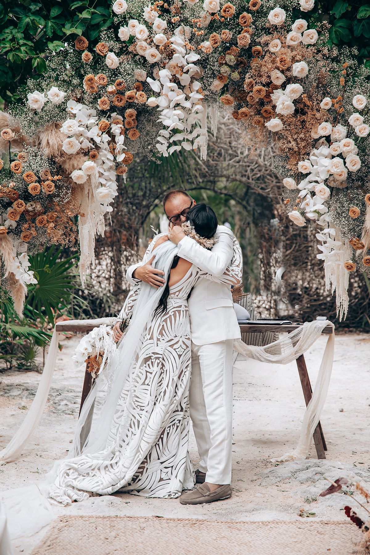 Romantic wedding portrait under a floral arch with white and peach roses against a natural backdrop.