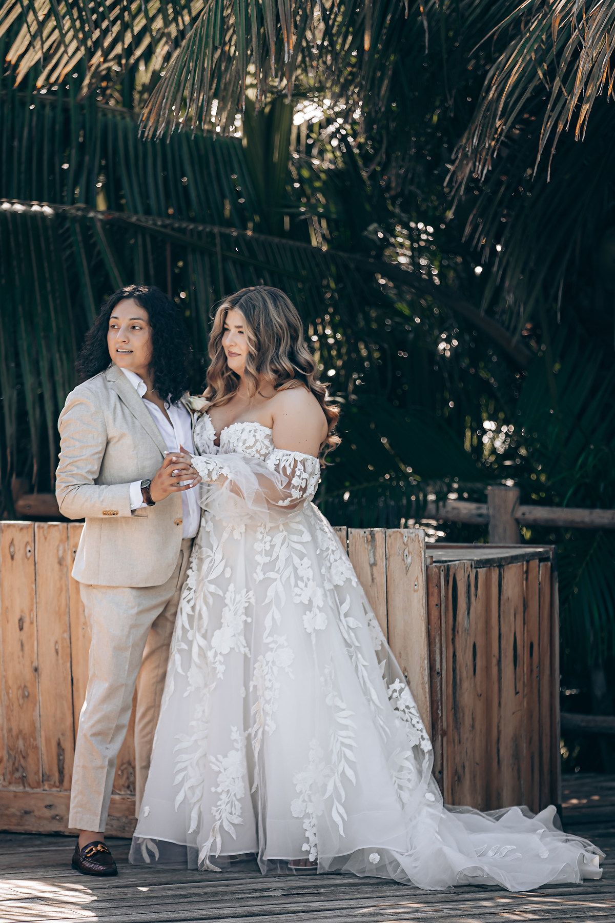 Two people in wedding attire share a tender moment on a rustic wooden bench surrounded by palm trees.
