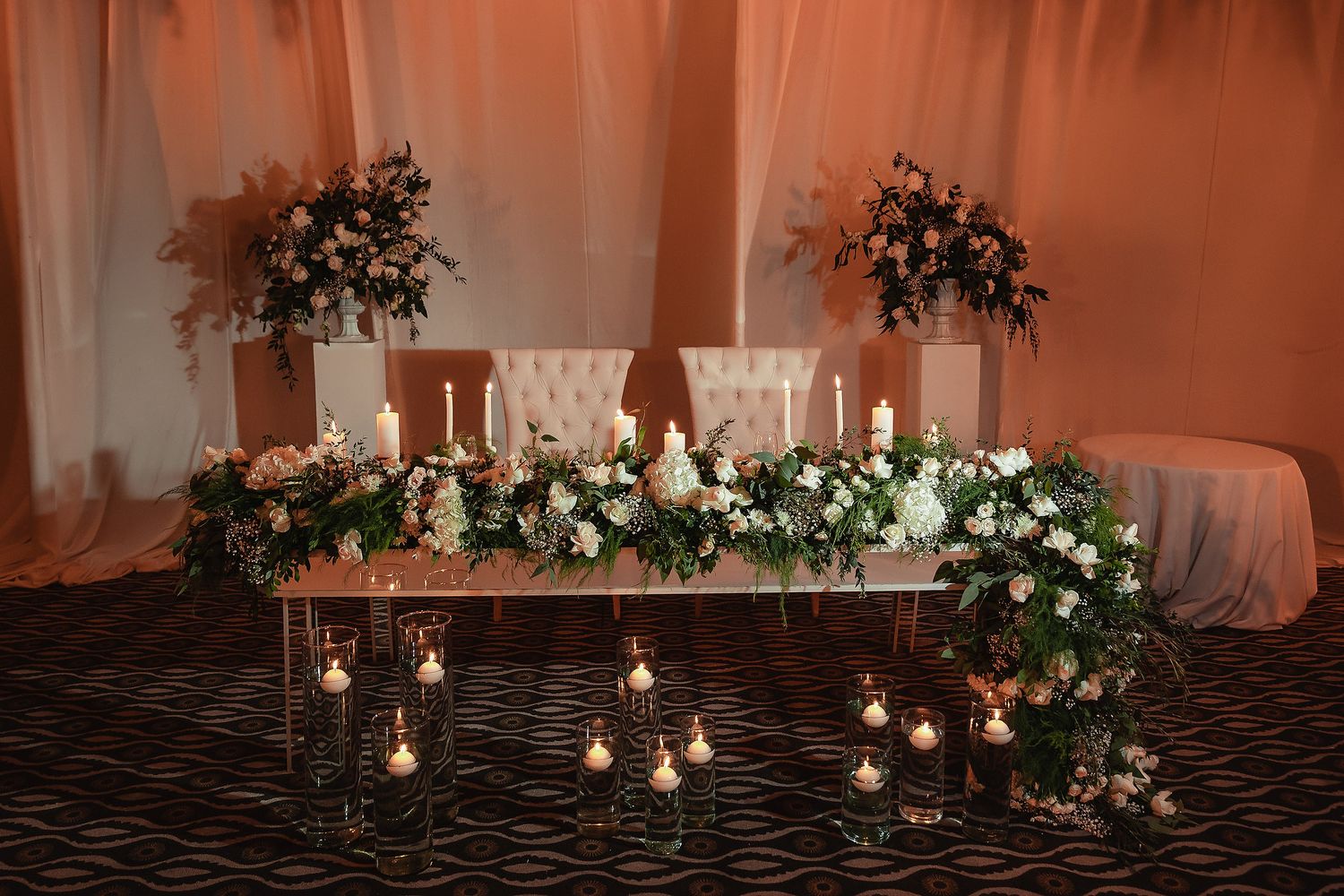 Elegant wedding reception decor with glowing candles, draped white fabric, and cascading greenery arrangements.