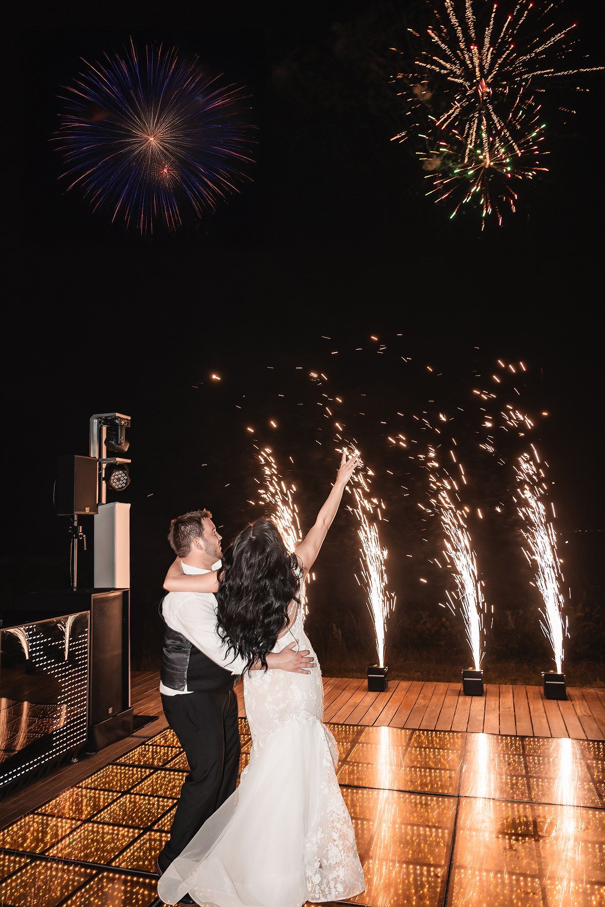 A couple dances on a wooden deck as fireworks burst in the night sky during their wedding celebration.