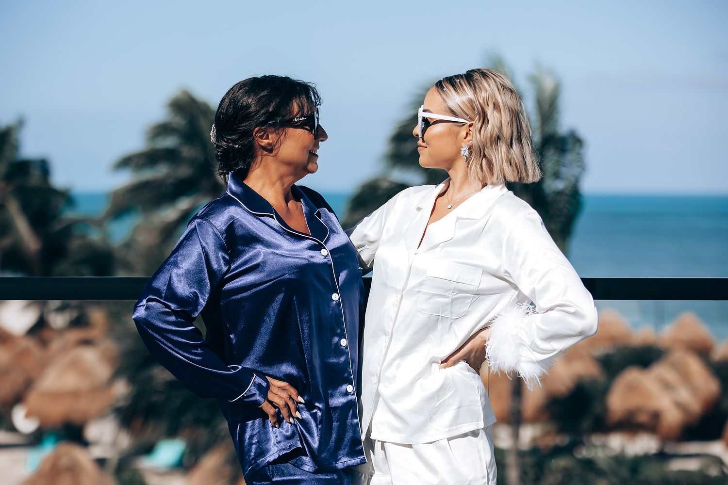 Two people in contrasting navy and white outfits dance together on a beach with ocean and rocks in the background.