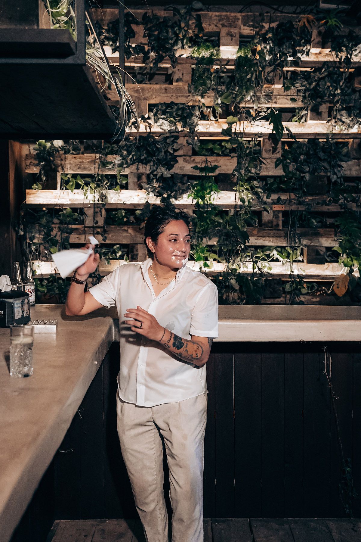 Person in white shirt and khaki pants poses against dark wood wall with hanging greenery in sequence of dance moves.