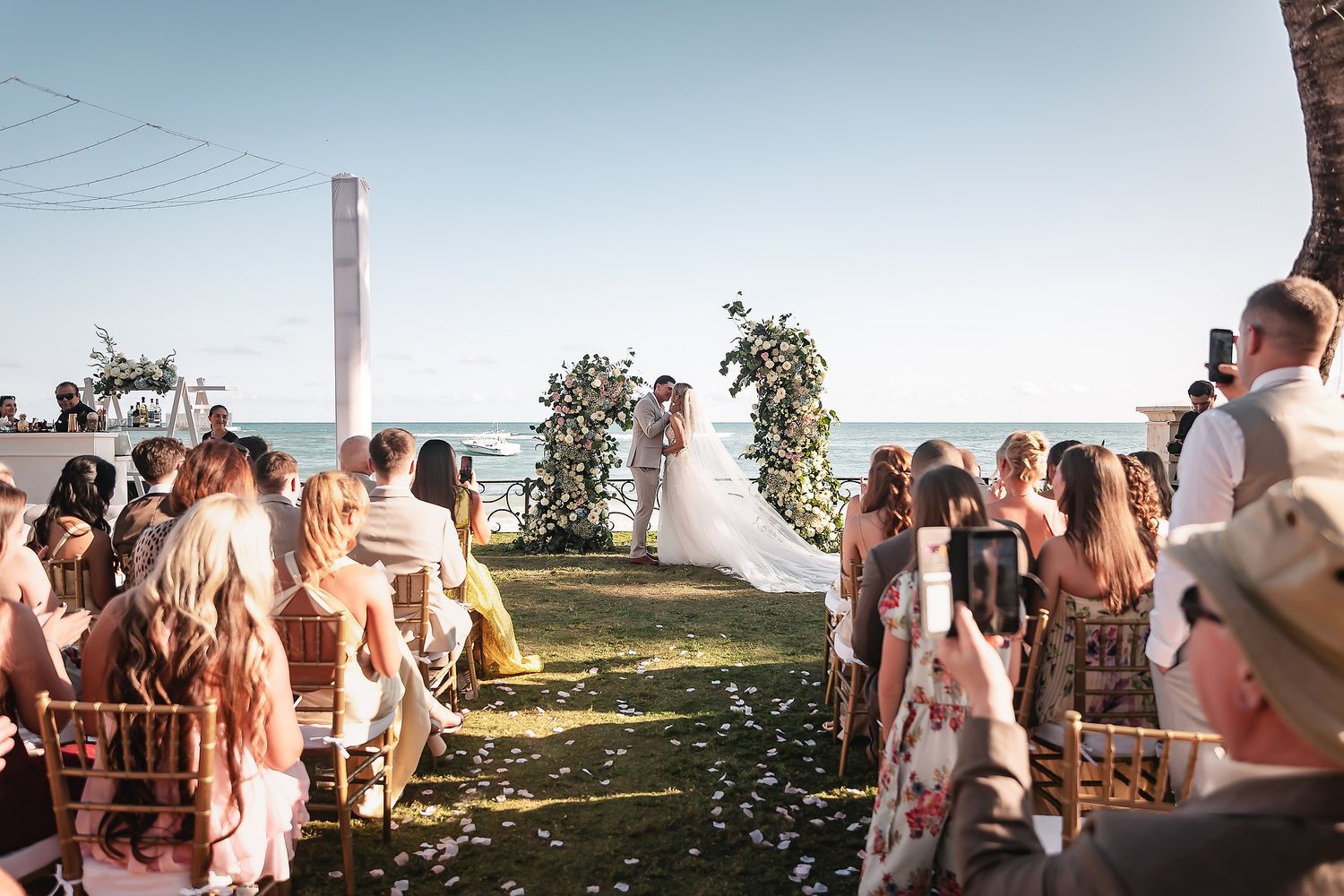 Wedding guests celebrate on a beach during sunset throwing flower petals in joyful celebration.