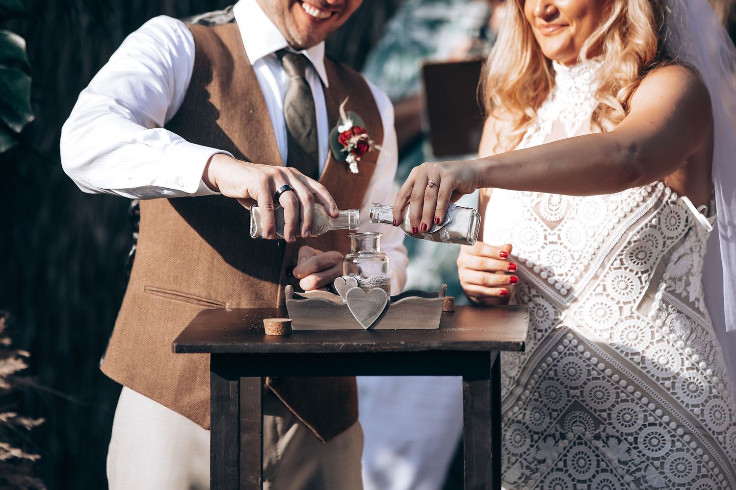 Two people perform a unity sand ceremony pouring colored sand into a container during an outdoor wedding celebration.
