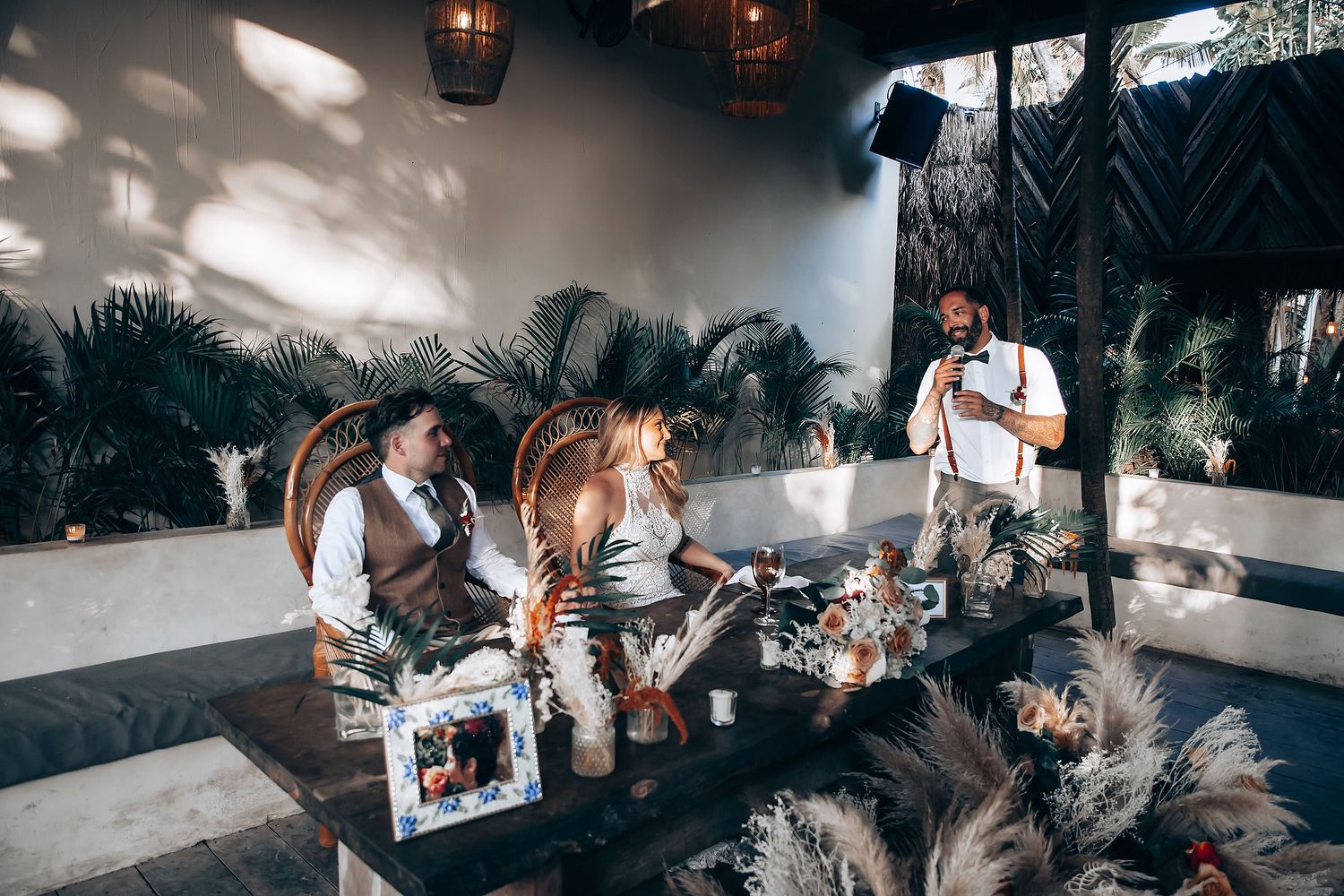 People gather at a tropical outdoor lounge area with pampas grass decor and hanging lanterns during a social event.