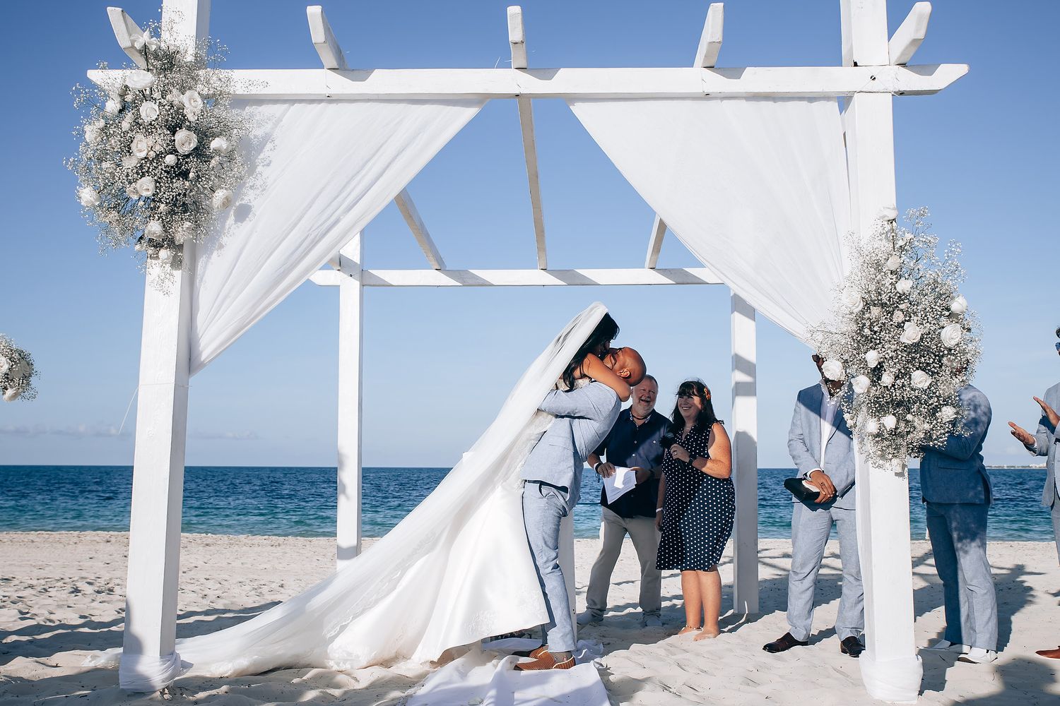 Elegant beach wedding setup with flowing white fabric and wooden pergola against blue ocean backdrop.