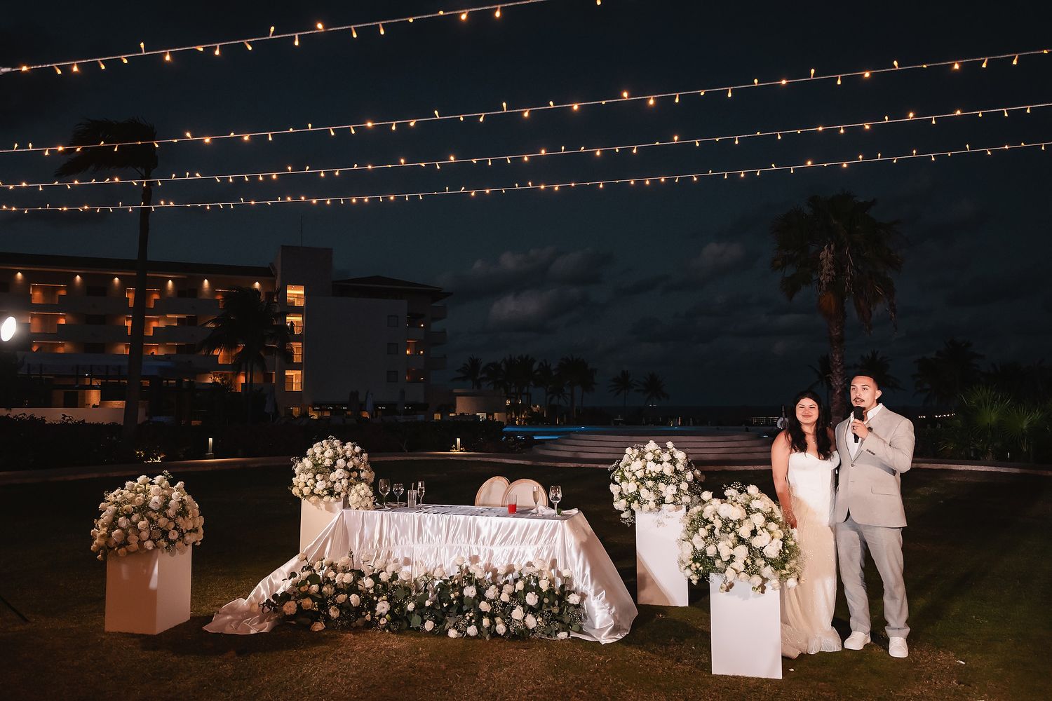 Evening wedding reception setup with string lights and floral arrangements on a lawn.