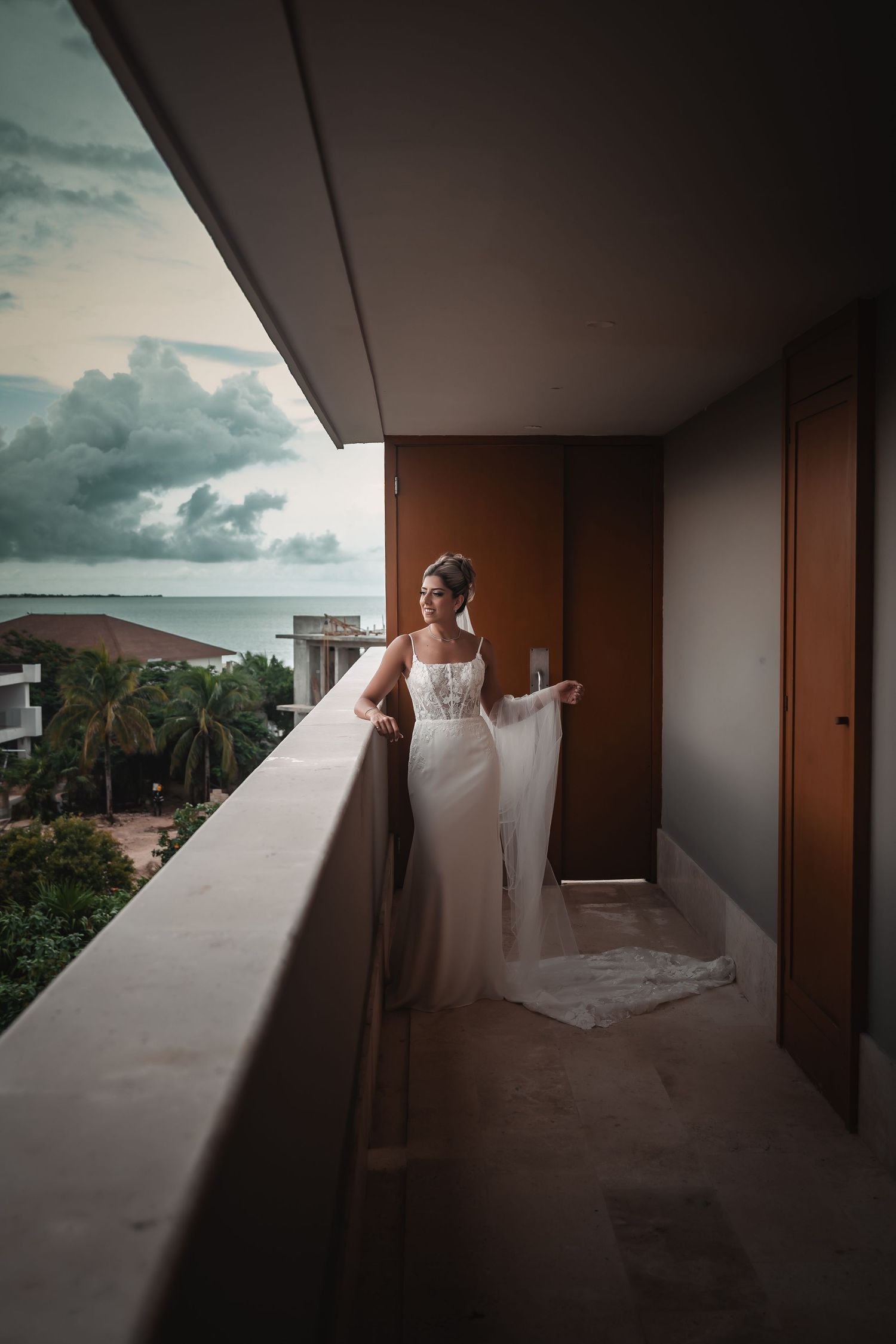 A bride in flowing white dress poses on a balcony against a dramatic cloudy sky.